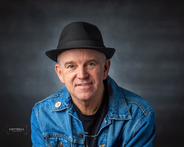 chris cotrell smiling and posing for a headshot in a black fedora and jean jacket