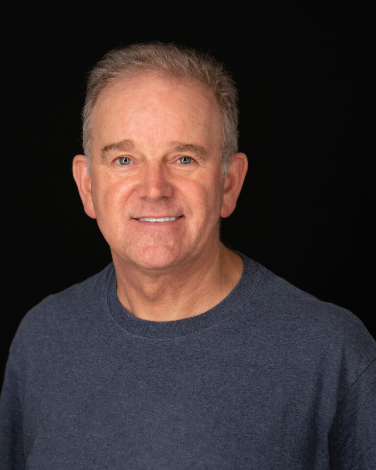 Chris wearing a dark blue tee shirt and looking at camera for portrait. Set against a black background under studio lighting.