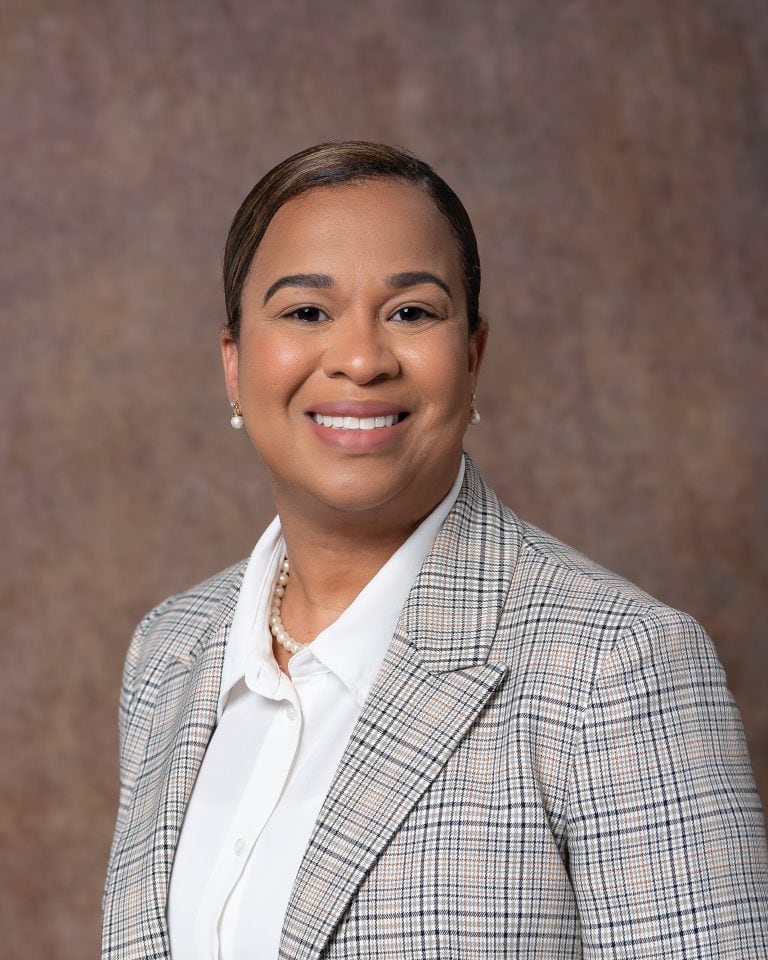 Female medical executive with short, dark-brown hair smiling at lens for professional headshots. Wearing a white top, gray, brown and beige blazer set agsinta textured brown, gold and light-brown backdrop.