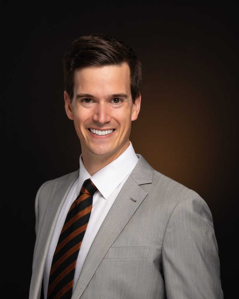 CEO smiling and looking at camera for professional headshot. Wearing a light grey suit, white dress shirt and dark brown and orange tie. Set against smooth, black backdrop, with an orange light behind which picks up the subjects eye color and tie. 
