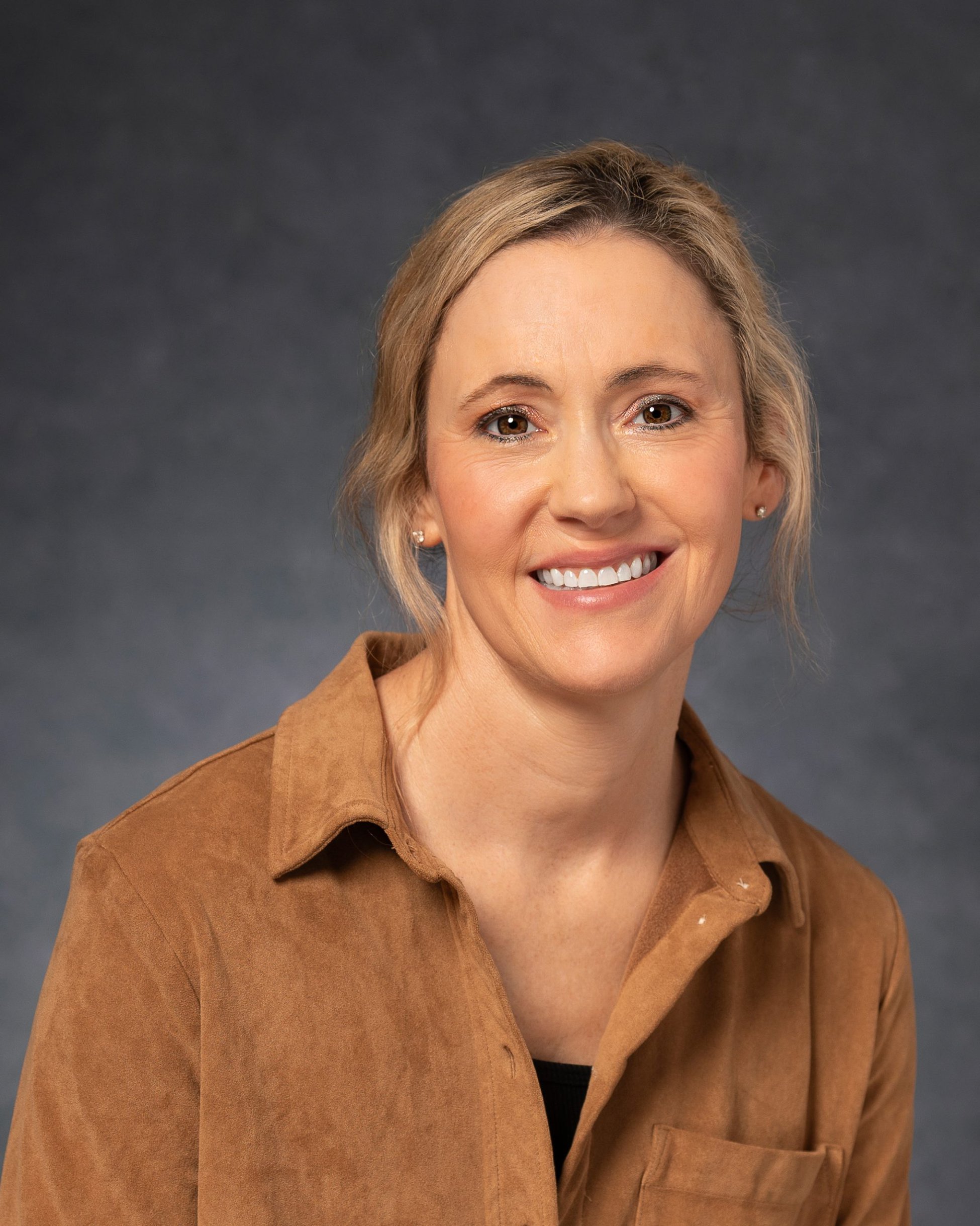 Small business owner smiling with a slight, empathetic tilt to her head. With short, dark blonde hair and brown eyes. Wearing black top and brown jacket set against a grey textured backdrop. 