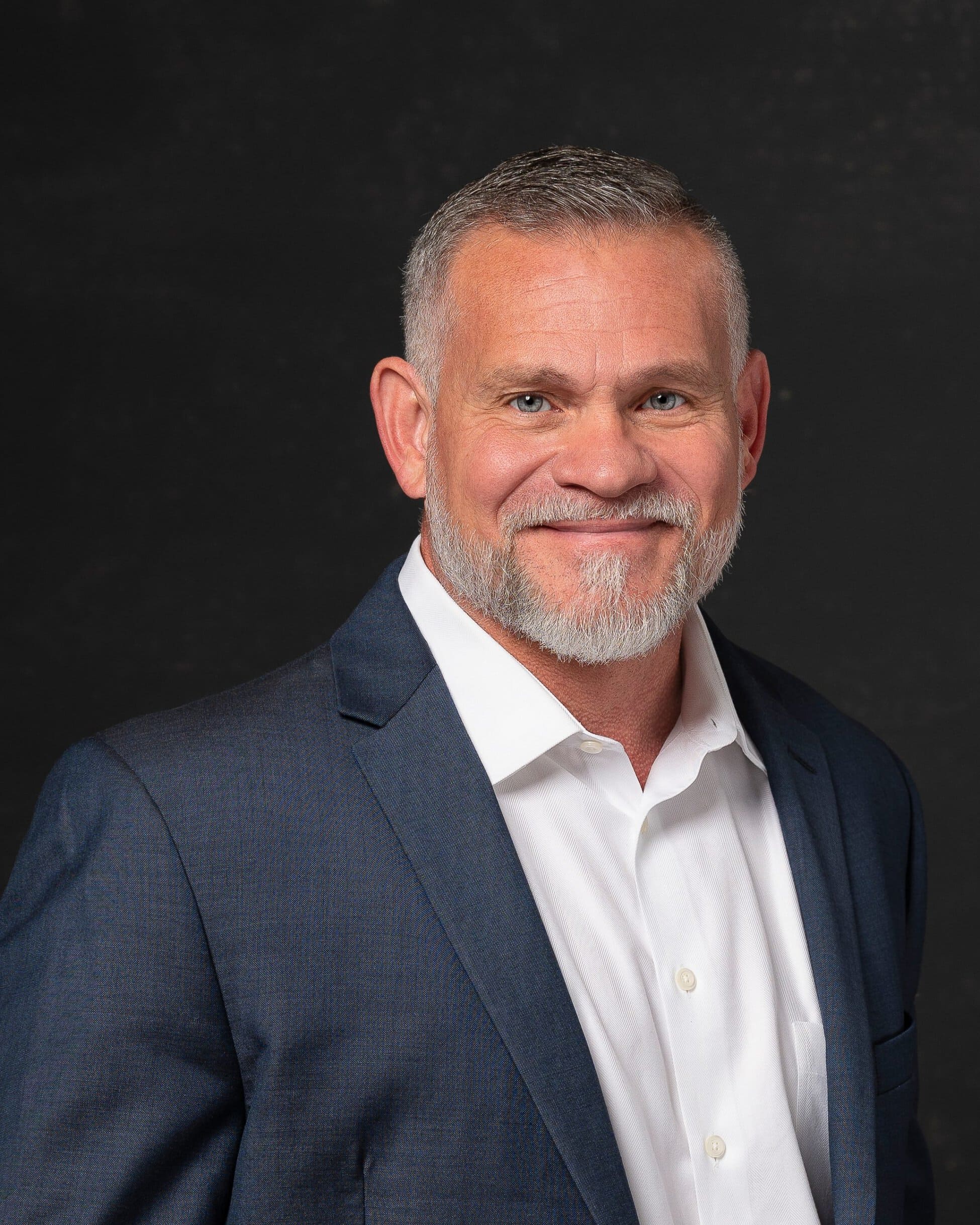 Construction industry executive with a grey beard, grey hair and bright blue eyes smiling while looking at camera for professional headshots. Wearing an open-collar white dress shirt and dark blue jacket. Shot under studio lighting and set against a dark grey backdrop. 
