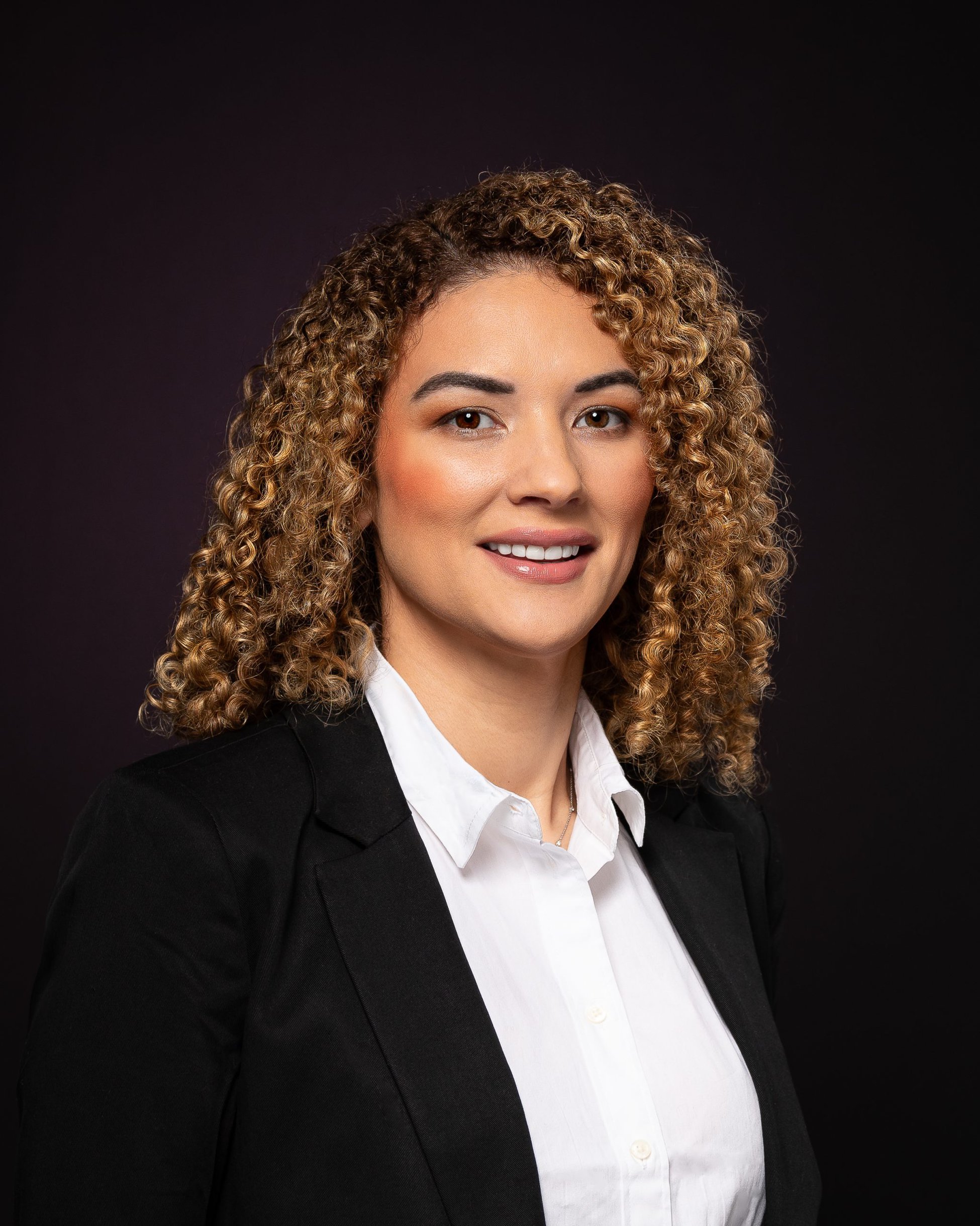 Business executive smiling while looking at camera for professional headshots. With light brown, hi lighted and curly hair and dark brown eyes. Wearing whit top and black blazer set against a black backdrop and shot under studio lighting. 