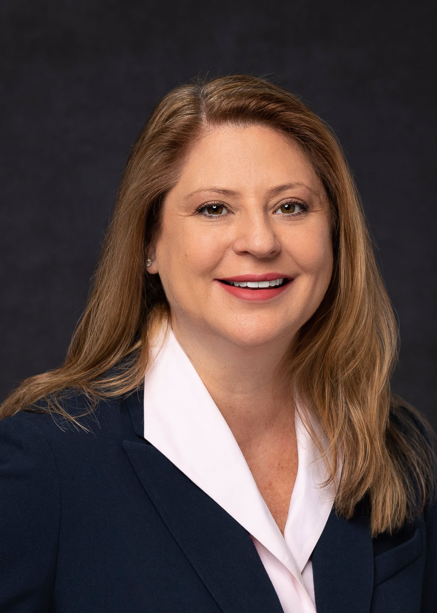 Insurance industry female executive smiling and looking directly at the lens for a professional headshot. With long, light brown and blonde hair and red lipstick. Wearing white top and dark blue blazer set against a dark-grey backdrop under studio lighting. 