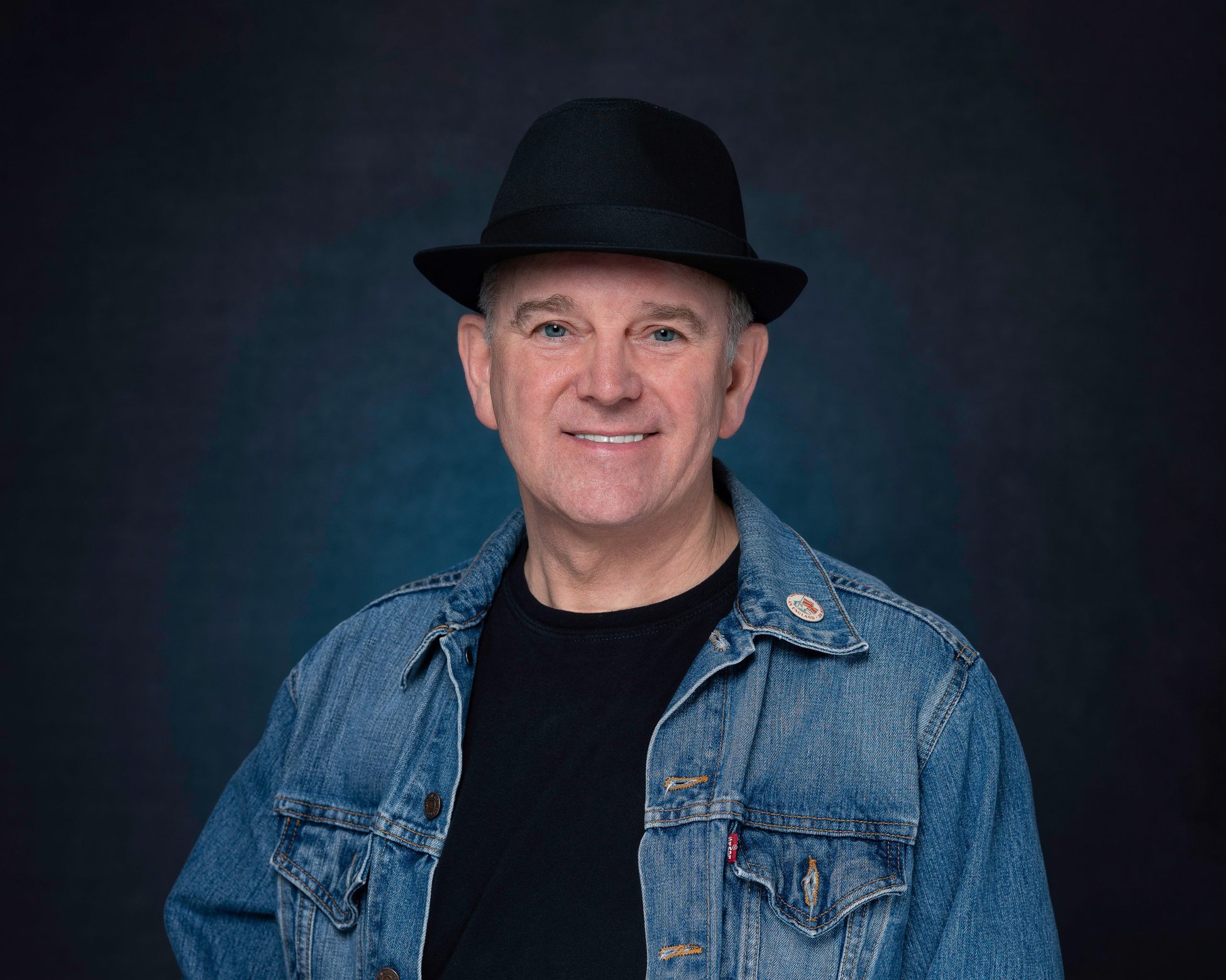 Professional Headshot Photographer Chris Cottrell looking directly at the camera lens for a headshot. Wearing his signature black fedora style hat, black tee shirt and denim jacket. Set against a grey, textured backdrop with added blue light behind. 