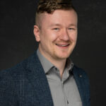 Professional headshot of man in gray collared shirt with friendly expression on black background