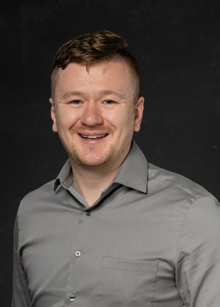 Professional headshot of man in light gray collared shirt against black background