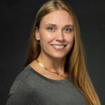Professional headshot of blonde woman in black blazer with warm smile against black background