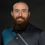 Professional headshot of bearded man in dark collared shirt with serious expression on black background