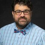 Professional headshot of man with glasses in light blue checkered shirt and bow tie against black background