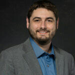 Professional headshot of bearded man in gray suit jacket and blue dress shirt against black background.