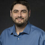 Professional headshot of bearded man in blue collared shirt smiling confidently on black background