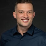 Professional headshot of smiling young man in dark navy collared shirt against black background.